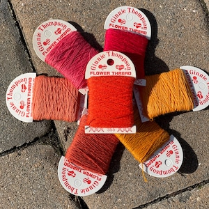 May include: Six spools of red, orange, and yellow embroidery floss with white labels that read "Ginnie Thompson Flower Thread".