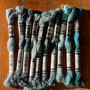 May include: A collection of DMC embroidery floss in various shades of blue and green. The spools are arranged on a wooden surface, showcasing a range of colors from light teal to deep turquoise. Each spool has a label with a number.