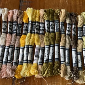 Puede incluir: Una colección de hilos de bordar DMC en varios tonos de blanco, amarillo y marrón. Las bobinas están etiquetadas con códigos de color y están dispuestas sobre una superficie de madera. Ideal para proyectos de punto de cruz y bordado.