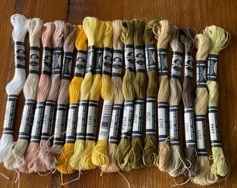 May include: A collection of DMC embroidery floss in various shades of white, yellow, and brown. The spools are labeled with color codes and are arranged on a wooden surface. Ideal for cross-stitch and embroidery projects.