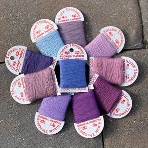 Può includere: Nove bobine di filo da ricamo in varie tonalità di viola, rosa e blu. Ogni bobina è etichettata "Ginnie Thompson Flower Thread 100% Cotone Made in USA".