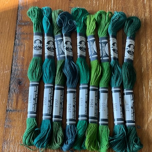 May include: Ten skeins of DMC embroidery floss in shades of green are displayed. Each skein has a label with a colour code and brand details. The floss is arranged on a wooden surface, highlighting the different tones of green.