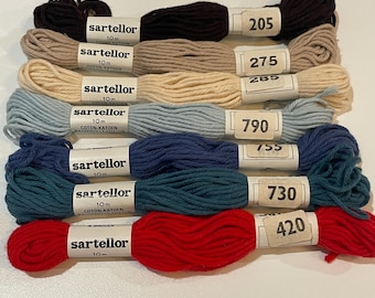 May include: A selection of colorful embroidery floss skeins. The floss is in shades of black, tan, cream, blue, teal, and red. Each skein has a paper label with the brand name and color number. The skeins are arranged horizontally.