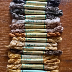 Puede incluir: Vista cenital de una colección de hilos de bordar de seda en varios tonos de gris, beige y amarillo. Cada madeja está etiquetada con la marca "Kreinik" e información sobre el contenido de seda y las instrucciones de cuidado.