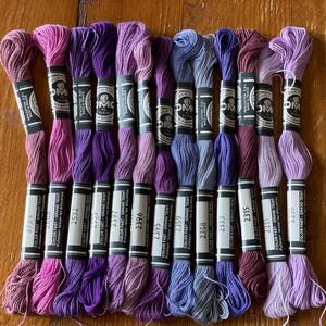 Pode incluir: Uma coleção de fios de bordar DMC em várias tonalidades de roxo, rosa e azul. Cada meada é rotulada com um número e as palavras "Color Fast Grand Teint". Os fios estão dispostos sobre uma superfície de madeira.