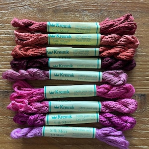 May include: A collection of silk embroidery floss skeins in various shades of pink and purple. Each skein is labeled "Kreinik Silk Mori" and "100% pure silk". The skeins are arranged on a wooden surface.