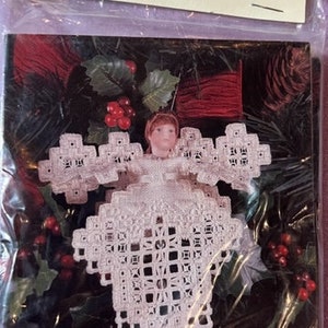 May include: A white cross stitch angel ornament with a red berry and holly leaf background. The angel is wearing a white dress and has a white halo. The ornament is packaged in a clear plastic bag with a white card that says "Cross 'N Patch" and "Genie Bishop".