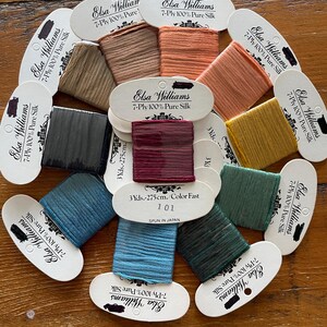 May include: A selection of Elsa Williams 7-Ply 100% Pure Silk threads in a variety of colours. The spools, each 275cm long, include shades of teal, green, and burgundy. Each spool is labeled with the brand name and product details. The threads are spun in Japan.
