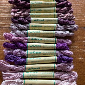 May include: A collection of silk embroidery floss in various shades of purple, from deep plum to pale lavender. Each spool is labeled with "Kreinik Silk Mori" and details about the silk content and care instructions. The spools are arranged neatly on a wooden surface.