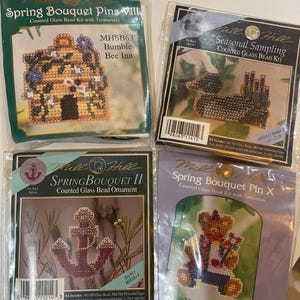 May include: Four Mill Hill counted glass bead kits in clear plastic packaging. The kits feature a yellow bumble bee inn, a brown duck, a pink anchor, and a teddy bear in a sailor outfit. Each package displays the kit name, model number, and contents.