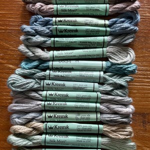 May include: A collection of Kreinik Silk Mori thread spools in various muted colors, including blues, greens, and browns. Each spool is labeled with the brand name, thread type, and care instructions. The spools are arranged on a wooden surface.
