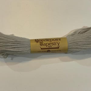 May include: A skein of light gray needlepoint tapestry yarn. The yarn is wrapped around a tan label with the words "Needlepoint Tapestry Yarn" in a decorative font. The label also states "100% Pure Virgin Wool".