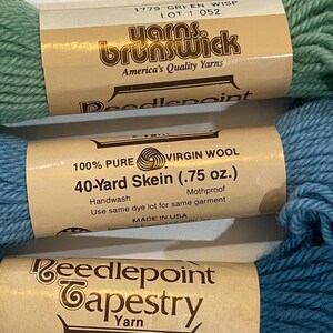 May include: Close-up of three skeins of yarn in shades of green and blue. The labels read "Yarns Brunswick" and "Needlepoint Tapestry Yarn." The blue skein is labeled "40-Yard Skein (.75 oz.)" and "100% Pure Virgin Wool."