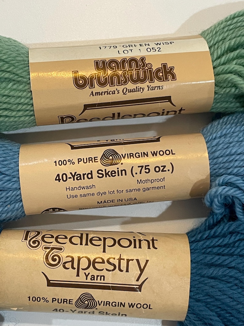 May include: Close-up of three skeins of yarn in shades of green and blue. The labels read "Yarns Brunswick" and "Needlepoint Tapestry Yarn." The blue skein is labeled "40-Yard Skein (.75 oz.)" and "100% Pure Virgin Wool."