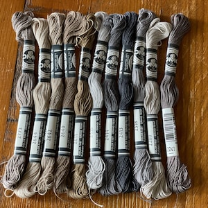 May include: A selection of DMC embroidery threads in a range of neutral tones, including browns, greys, and beiges. Each skein is labelled with a colour number and brand details. The threads are arranged on a wooden surface, perfect for embroidery or cross-stitch.