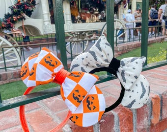May include: Two Halloween-themed headbands. One has an orange and white checkered bow with jack-o'-lantern faces. The other has a white bow with gray bats. Both have black headbands. The first headband has an orange velvet band.