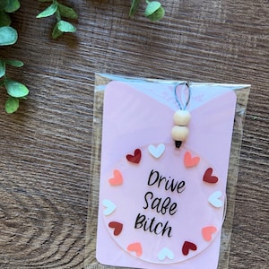 May include: A pink card with a clear round charm that says "Drive Safe Bitch" in black lettering. The charm is surrounded by pink, white, and red hearts. The card has a white string with two wooden beads attached.