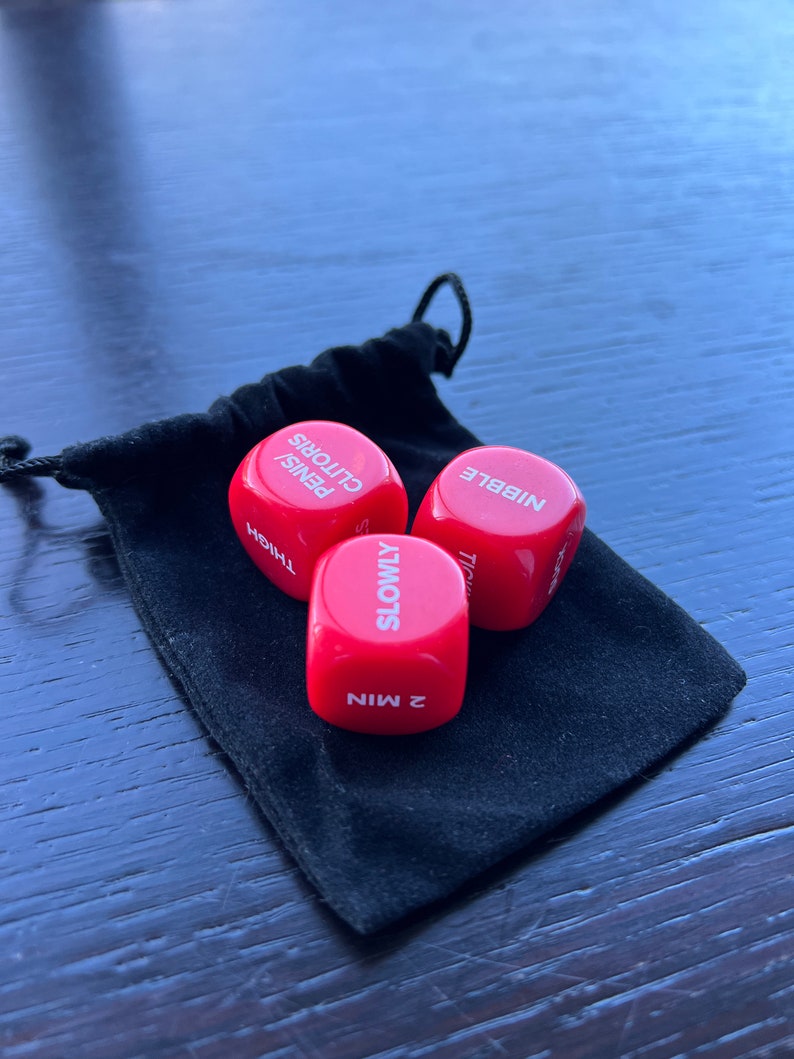 May include: Three red dice with white text. The dice read "2 MIN", "SLOWLY", "THIGH", "PENIS/CLITORIS", "NIBBLE". The dice are in a black velvet bag.