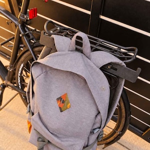 May include: A heather gray backpack with a top handle, adjustable shoulder straps, and a front pocket. The bag features a diamond-shaped patch with an orange and green camouflage pattern and a white rectangular label with the Herschel brand name.