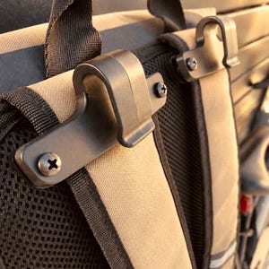 May include: A tan and black backpack featuring two black plastic J-shaped hooks attached to the top of the shoulder straps with metal screws. The backpack has a black mesh back panel and a black nylon top handle, designed for secure attachment.