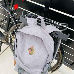 May include: A gray backpack with a camouflage patch and a small front pocket is attached to a black bicycle. The backpack has a top handle and adjustable straps. The bottom of the backpack has a brown and green pattern.