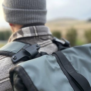 Może przedstawiać: Szara czapka i szara koszula w kratę noszone pod szałwiowo-zielonym plecakiem z czarnymi paskami i czarnym metalowym mocowaniem zabezpieczonym widocznymi śrubami na pasku naramiennym, z rolowanym zamknięciem i czarnymi nylonowymi taśmami.