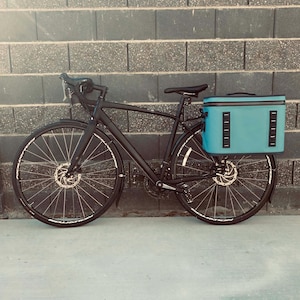 Könnte beinhalten: Ein schwarzes Fahrrad mit einer türkisblauen Aufbewahrungstasche am Heck. Das Fahrrad hat Scheibenbremsen und ein modernes Design. Der Hintergrund ist eine graue Backsteinmauer.