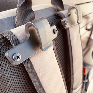 May include: Close-up of a grey and black backpack with metal clips. The clips are attached with screws and are designed to secure the backpack's contents. The backpack has a mesh back panel and grey straps.