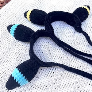 Pode incluir: Duas faixas de cabeça de crochê pretas com detalhes em azul e amarelo. As faixas de cabeça estão sobre uma superfície de crochê branca.