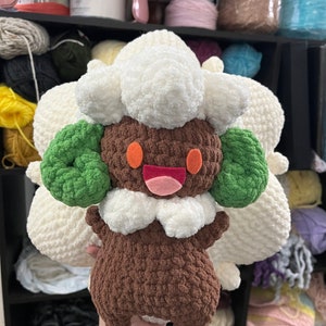 May include: A crocheted plush toy of a brown and white Pokemon character with green ears. The toy is being held by a hand.
