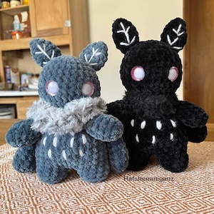 May include: Two handmade plush creatures. One is dark blue with a gray collar and white accents, the other is black with white accents. Both have large eyes with pink glitter and leaf-shaped ears. The plush toys are on a patterned surface.
