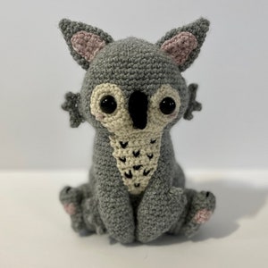 May include: A handmade gray crochet creature with large black eyes, a cream-colored face, and pink accents on the ears and feet. The amigurumi-style toy has small arms and is sitting down. A unique, whimsical stuffed animal.