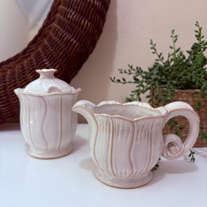 May include: A cream-colored ceramic sugar bowl with a lid and a matching creamer. Both pieces have a tulip-like design with vertical ridges and a scalloped edge. The creamer has a handle and a spout.