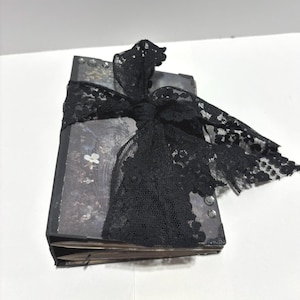 Puede incluir: Un diario rectangular con una cubierta oscura de estampado floral que presenta pequeñas flores blancas. El libro está atado con una cinta ancha de encaje negro anudada en la parte superior. Las esquinas están reforzadas con piezas metálicas.