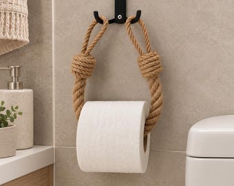 May include: A toilet paper holder featuring a thick, natural-colored jute rope looped through a black metal wall-mounted hook. The rope is secured with two prominent knots on either side, suspending a standard roll of white toilet paper.