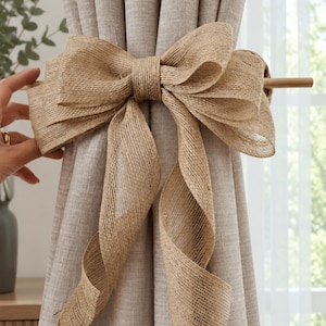 May include: A beige curtain tieback featuring a large, decorative bow made from textured, light brown burlap fabric. The bow is attached to a thin, horizontal wooden rod that secures the gathered fabric of the light beige curtain panel.