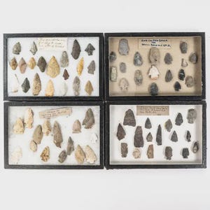 May include: Four framed displays of ancient arrowheads. The arrowheads are in various shapes and colors, including tan, gray, and brown. Each display has a label with handwritten text. The frames are black and the background is white.