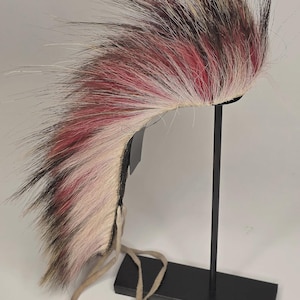 May include: A colorful, faux fur headdress with a black base and a long, flowing crest. The crest features shades of pink, white, and brown. The headdress is displayed on a black metal stand with a rectangular base and a beige strap.