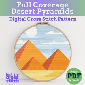 May include: A digital cross stitch pattern featuring a desert landscape with three orange pyramids under a blue sky with white clouds. The pattern is displayed on a wooden embroidery hoop.