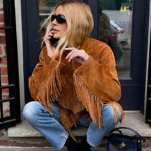 Peut inclure: Une veste en daim marron clair avec de longues franges sur la poitrine et les manches, associée à un jean bleu clair, des bottines noires et un sac à main en cuir bleu marine structuré avec un fermoir en métal doré.