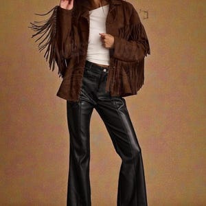 May include: A brown suede jacket with long fringe detailing on the sleeves and chest, worn over a white top and black leather-look flared trousers. The outfit includes black pointed-toe heels and dark sunglasses, with a West Crush logo in the corner.