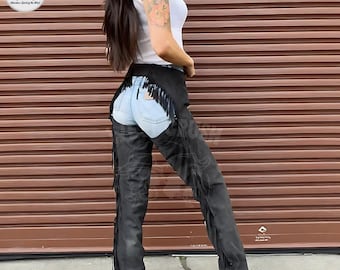 May include: Black leather chaps with fringe detailing. The chaps are worn over light blue denim jeans. The chaps have a decorative design on the legs. The model is wearing a white tank top.
