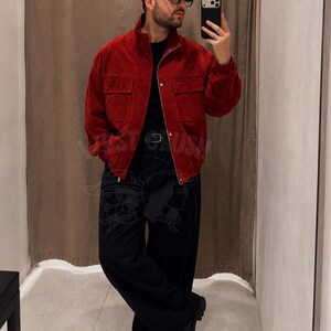 Puede incluir: Una chaqueta de ante rojo oscuro con cuello alto, cremallera frontal completa y dos grandes bolsillos en el pecho. La chaqueta se lleva sobre una camiseta negra lisa, combinada con pantalones negros holgados y botas de cuero negro.