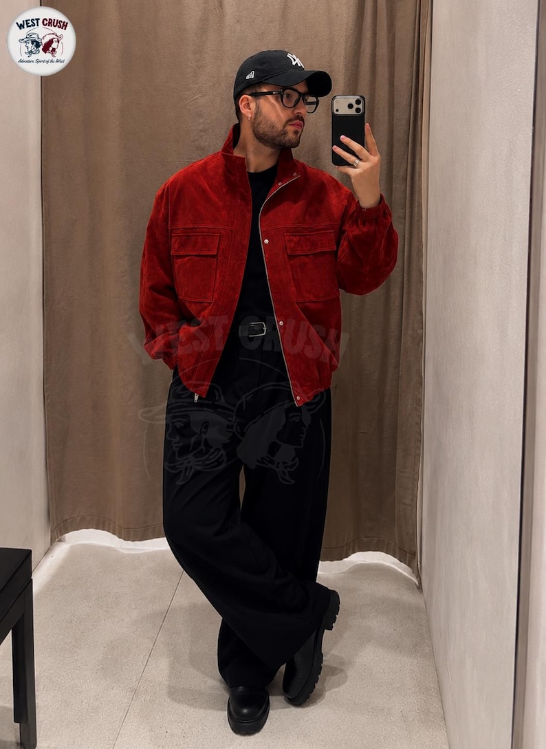 Puede incluir: Una chaqueta de ante rojo oscuro con cuello alto, cremallera frontal completa y dos grandes bolsillos en el pecho. La chaqueta se lleva sobre una camiseta negra lisa, combinada con pantalones negros holgados y botas de cuero negro.