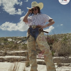 May include: A person wearing a straw cowboy hat, sunglasses, and a striped shirt, paired with denim jeans and chaps. The chaps feature a design with the text "ES CR". A belt with a decorative buckle completes the look, set against a backdrop of a blue sky with clouds.