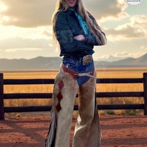 Puede incluir: Una mujer con sombrero de vaquero, vaqueros, camisa verde oscuro y chaps color canela con flecos. Los chaps tienen detalles decorativos rojos y marrones. Se ve una hebilla de cinturón. El fondo presenta una valla de madera y un paisaje con montañas.