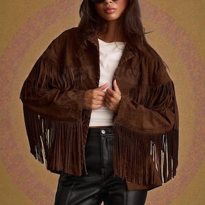 May include: A brown suede jacket with long fringe detailing on the sleeves and chest. The jacket is worn over a white top and paired with black leather trousers. The image features a logo in the upper left corner that says "West Crush".