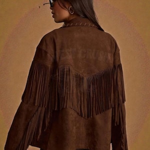 May include: A brown suede-textured jacket featuring long fringe detailing across the back and sleeves. The garment includes a collar with visible stitching and a V-shaped yoke design on the back. The text WEST CRUSH is embossed on the back.