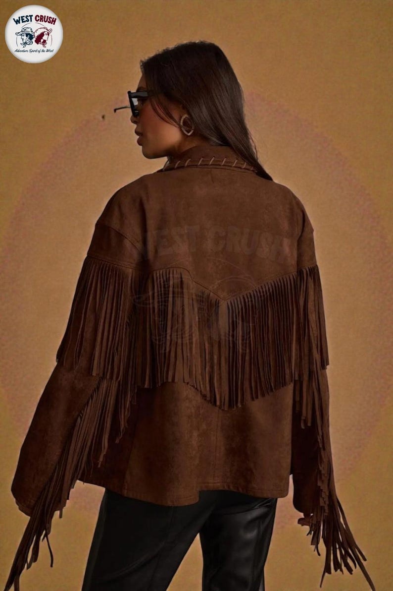 May include: A brown suede-textured jacket featuring long fringe detailing across the back and sleeves. The garment includes a collar with visible stitching and a V-shaped yoke design on the back. The text WEST CRUSH is embossed on the back.