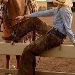 Puede incluir: Una persona con sombrero de vaquero, camisa a cuadros azul y chaps marrones, está sentada en una valla junto a un caballo. Los chaps tienen flecos. La imagen tiene una estética cálida al aire libre, con el texto "WEST CRUSH" visible.